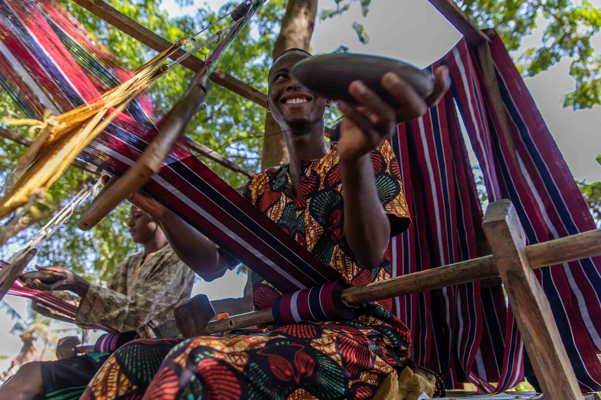 An aso-oke weaver at a wooden loom, holding up the shuttle